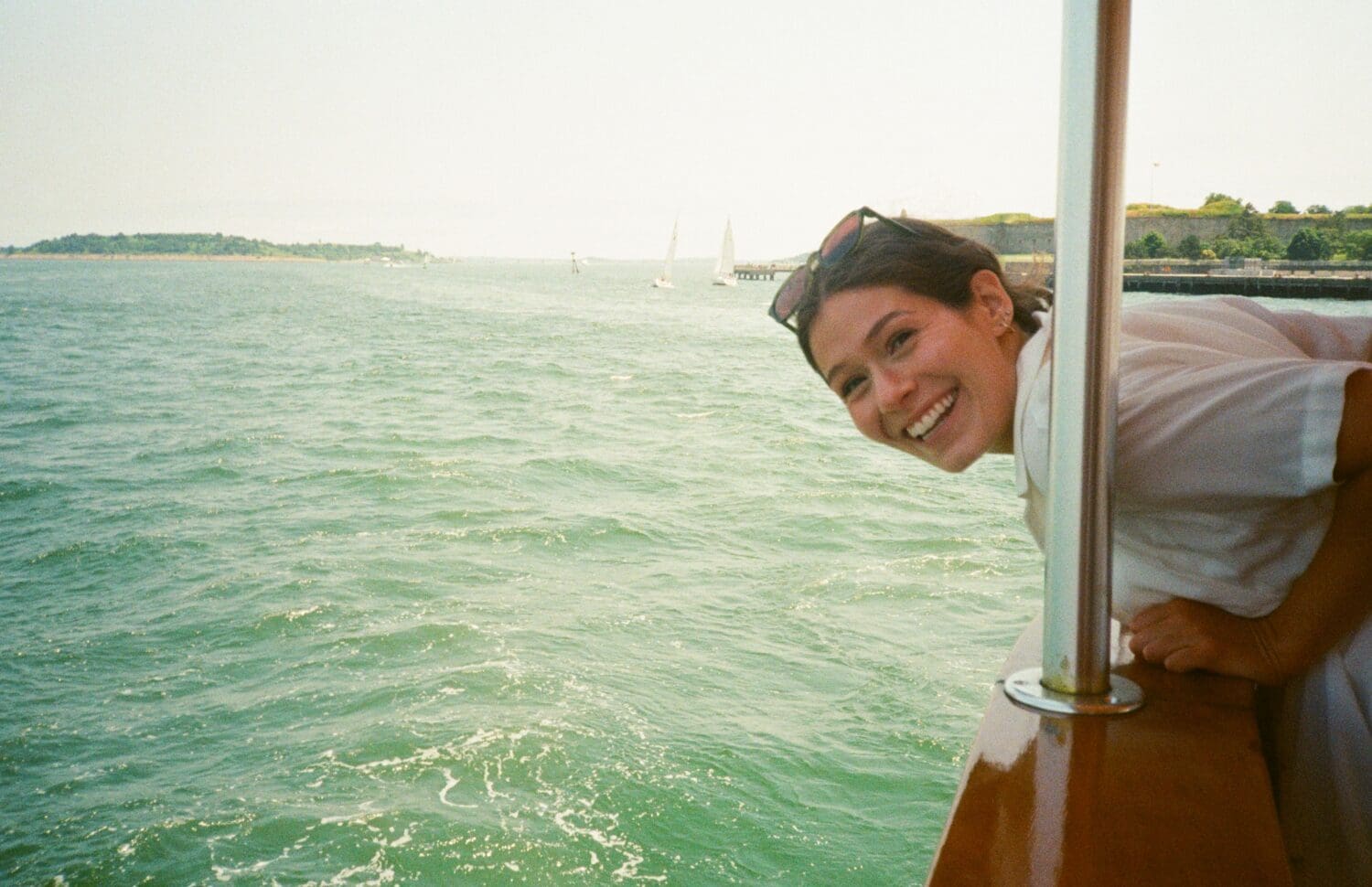 A person leans over the railing of a boat, smiling at the camera with a view of the sea and distant land in the background.