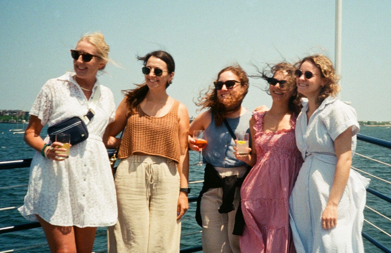Five women stand together on a boat, smiling, with drinks in hand, against a backdrop of water and a clear sky.