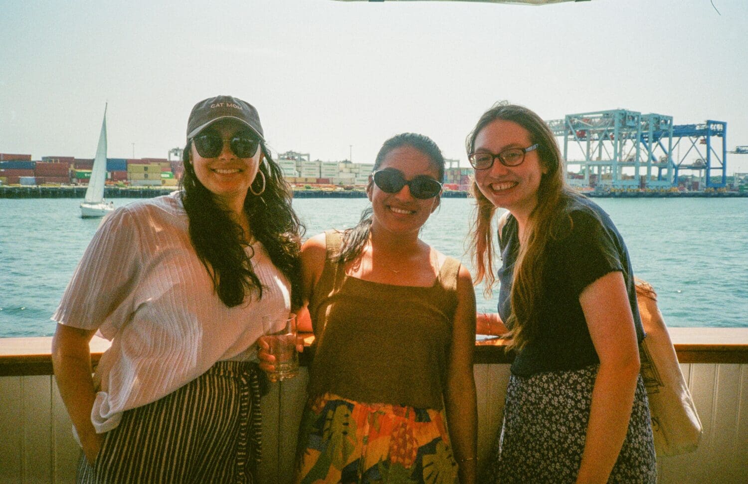 Three people smiling on a boat with water, a sailboat, and industrial structures in the background.