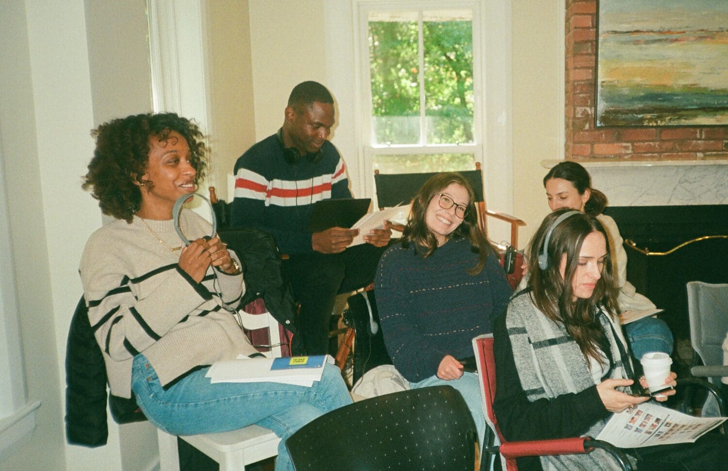 Five people are sitting in a room, some holding papers and a coffee cup, while conversing and smiling. A painting and a window are in the background.