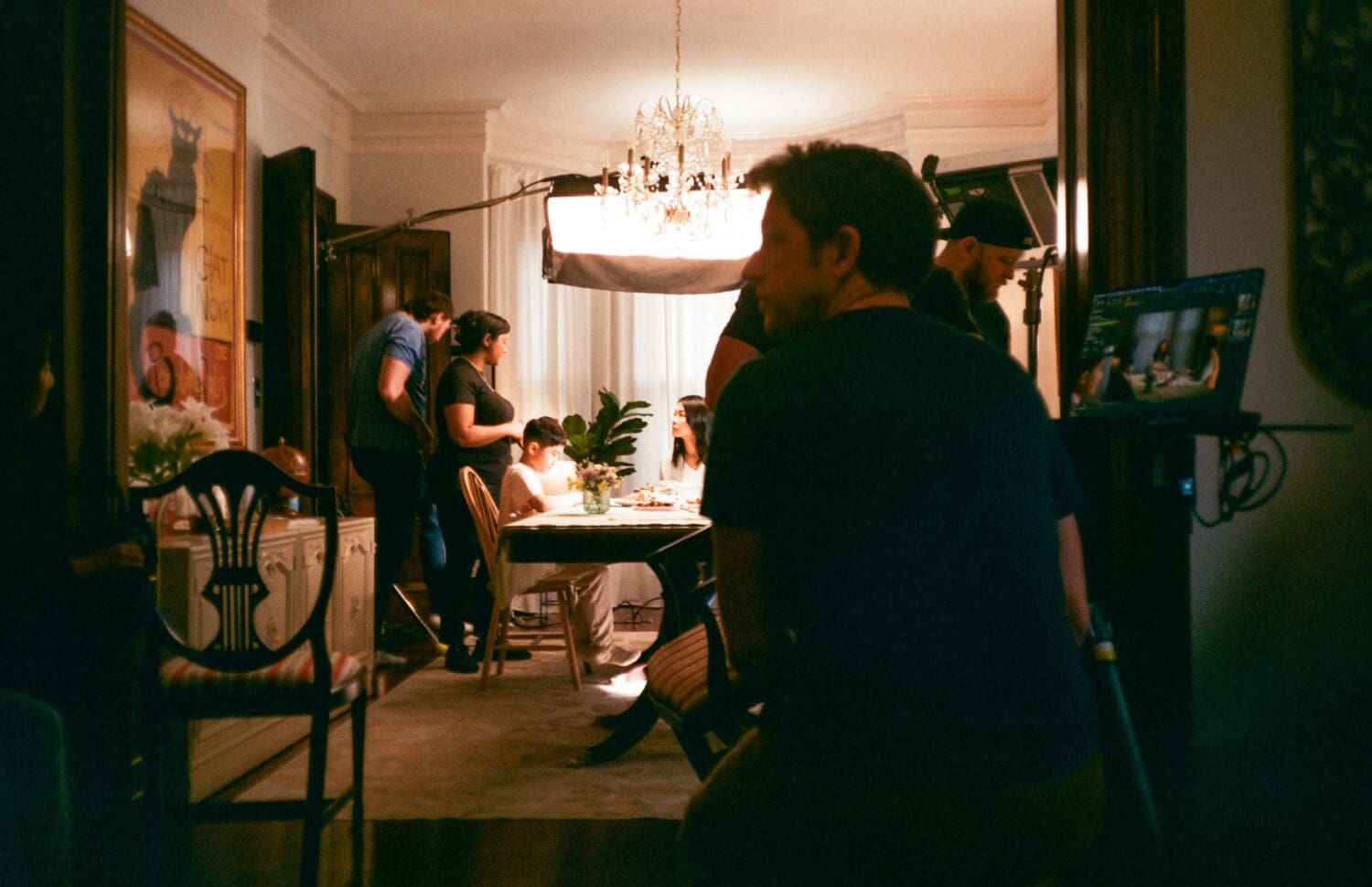 A film crew is setting up a dining room scene, with lights and camera equipment. Several people are around a table, and a chandelier hangs overhead.