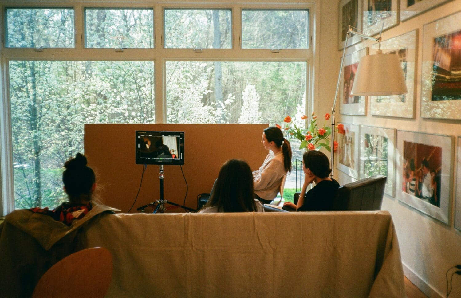 Four people seated on a couch watch a small screen in a bright room with large windows and framed pictures on the walls.