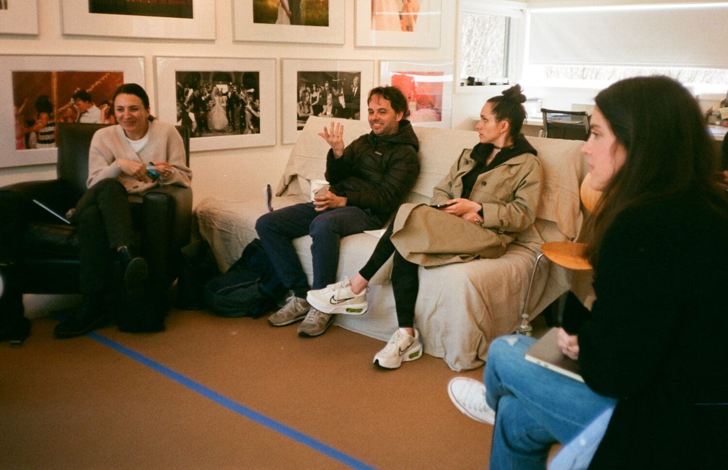 A group of people sitting on couches and chairs in a room with framed photos on the walls, engaged in conversation.