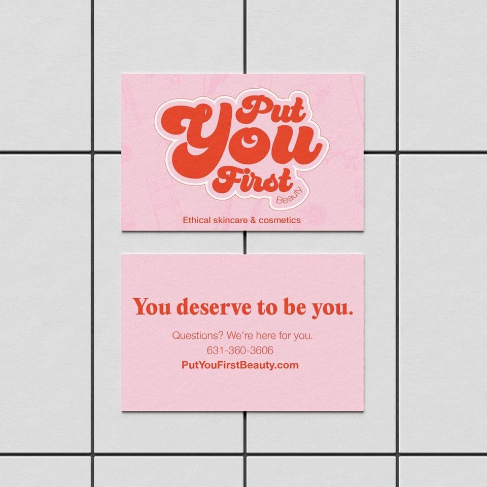 Two pink business cards for Put You First Beauty. One side has the brand name and slogan, the other displays a supportive message, contact number, and website on a tiled surface.