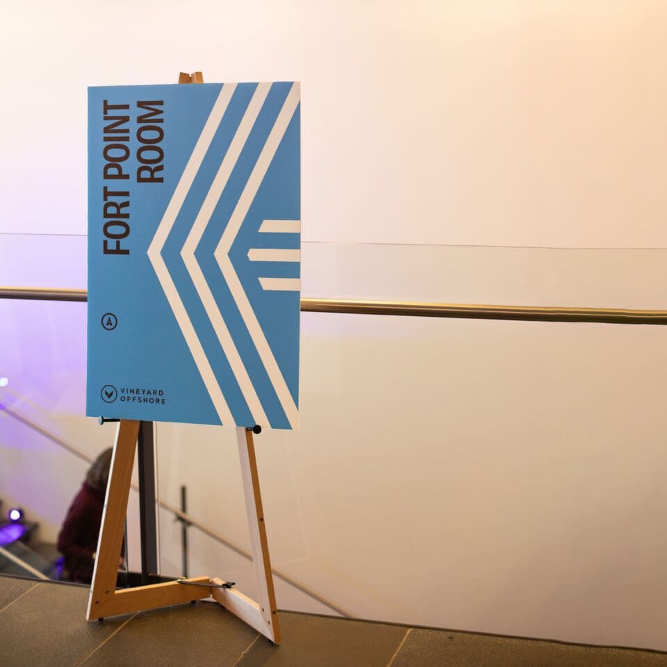Sign on an easel for "Fort Point Room" with blue and white geometric design, beside a glass railing.