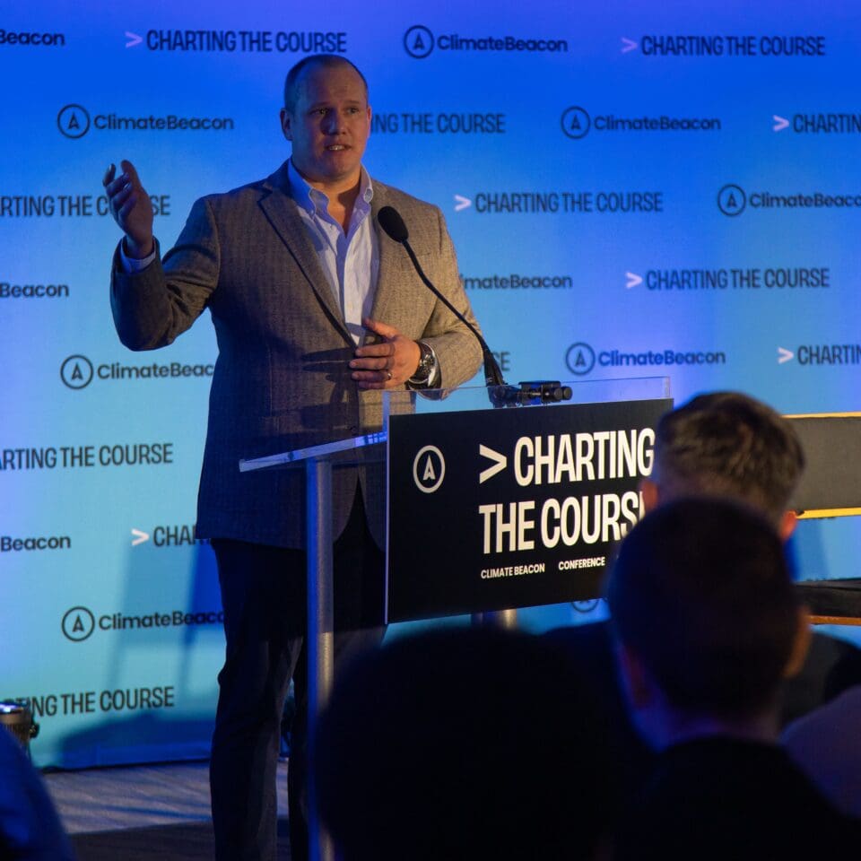 A person in a suit speaks at a podium during a "Charting the Course" event, with a blue backdrop featuring "Climate Beacon" logos.