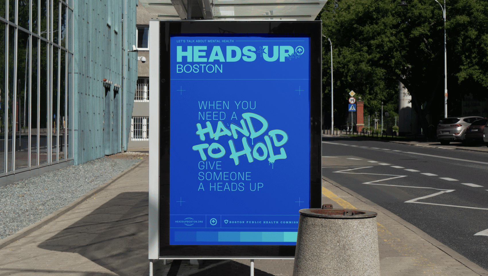 Bus stop ad with blue poster reading "When you need a hand to hold, give someone a heads up." Located on a sidewalk near a road with trees and buildings in the background.