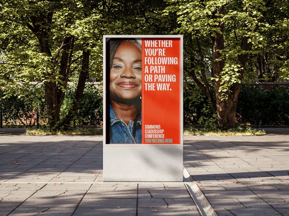 A street sign displays a woman's portrait beside text that reads: “Whether you’re following a path or paving the way. Simmons Leadership Conference. You belong here.”.