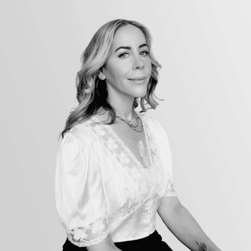 Woman with light wavy hair wearing a white blouse and necklace, sitting and facing the camera against a light gradient background.