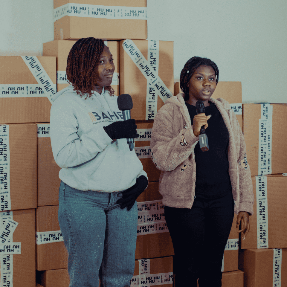 Two people holding microphones stand in front of stacked brown boxes.