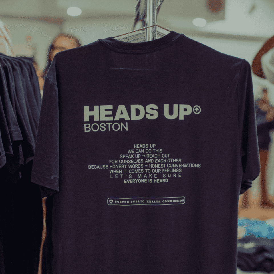 Black T-shirt on a rack with white text promoting mental health awareness in Boston, emphasizing communication and support.