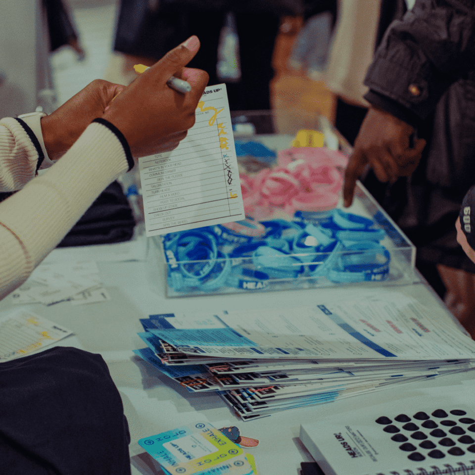 Person holding a form and pen near a table covered with documents, brochures, and wristbands during an event registration or information session.