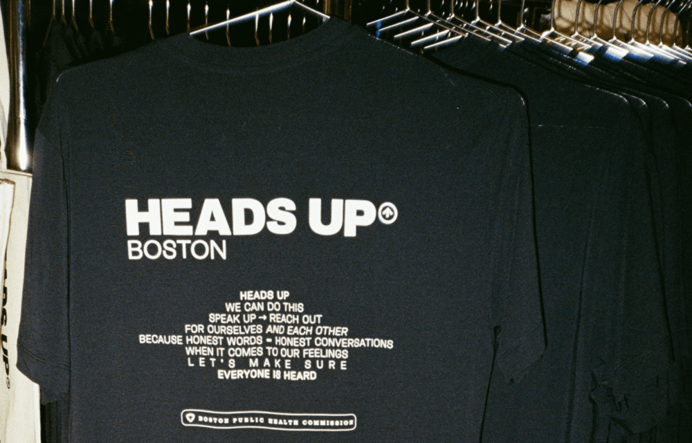 Navy blue t-shirt on a rack displaying "Heads Up Boston" and a message encouraging open conversations about feelings, issued by the Boston Public Health Commission.