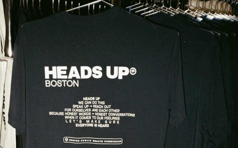 Navy blue t-shirt on a rack displaying "Heads Up Boston" and a message encouraging open conversations about feelings, issued by the Boston Public Health Commission.