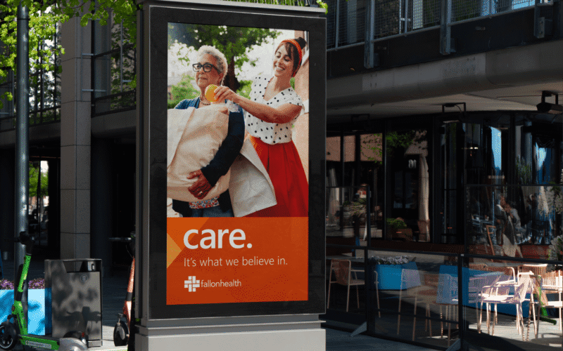 Outdoor advertisement featuring two people smiling and carrying laundry, with the text "care. It's what we believe in." and a logo for #allonhealth.