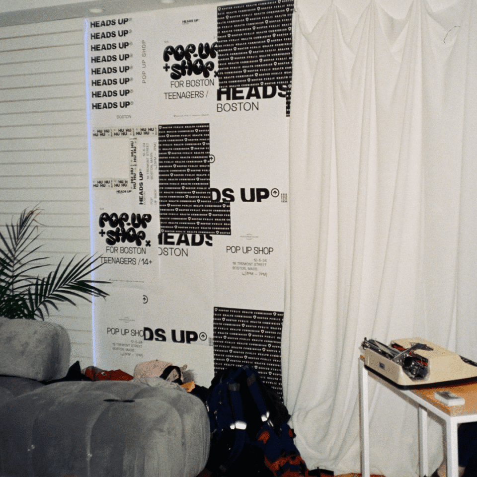 Room interior with a gray couch, indoor plant, scattered clothes, a bag, and a typewriter on a wooden table. The wall has posters for a "Pop Up Shop: Heads Up for Boston Teenagers.