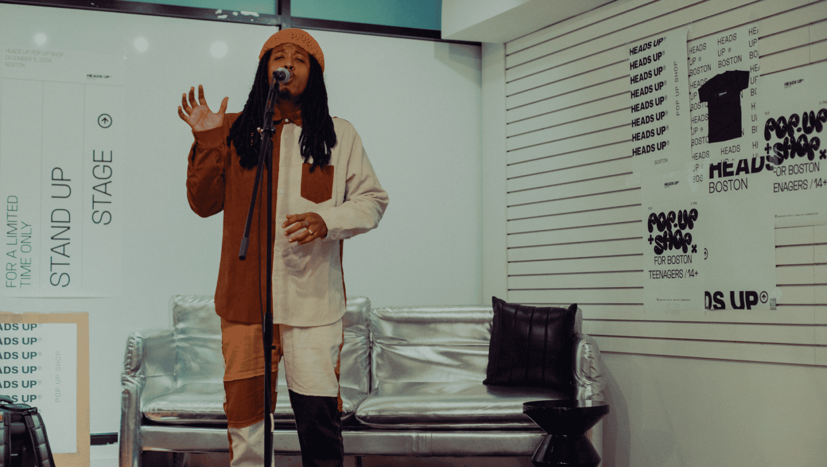 Person with dreadlocks and colorful outfit performs on stage with microphone, surrounded by modern decor and graphic posters.