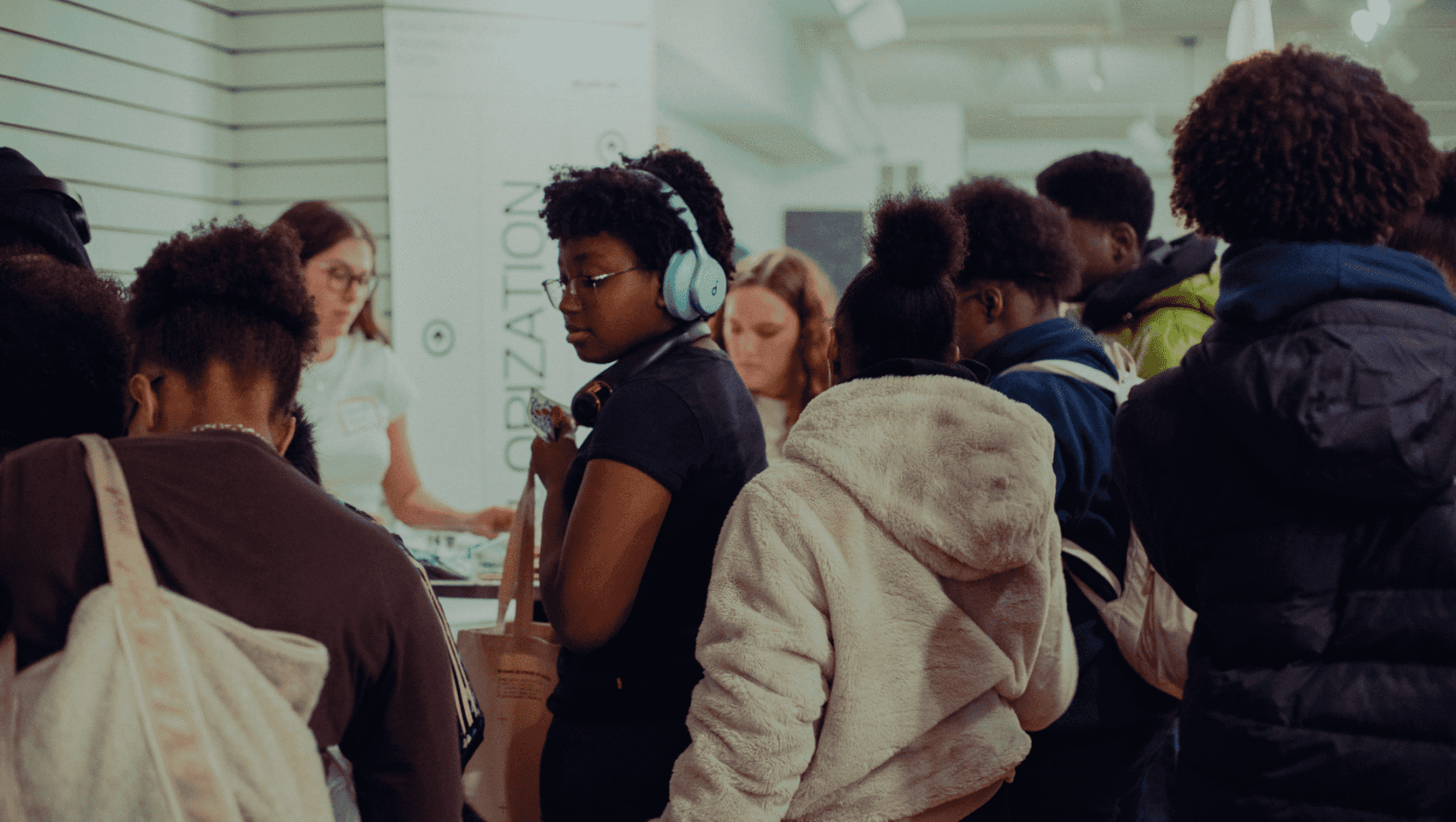 A group of people, some wearing backpacks and headphones, stand in a crowded indoor space, facing a counter where a woman is assisting them.