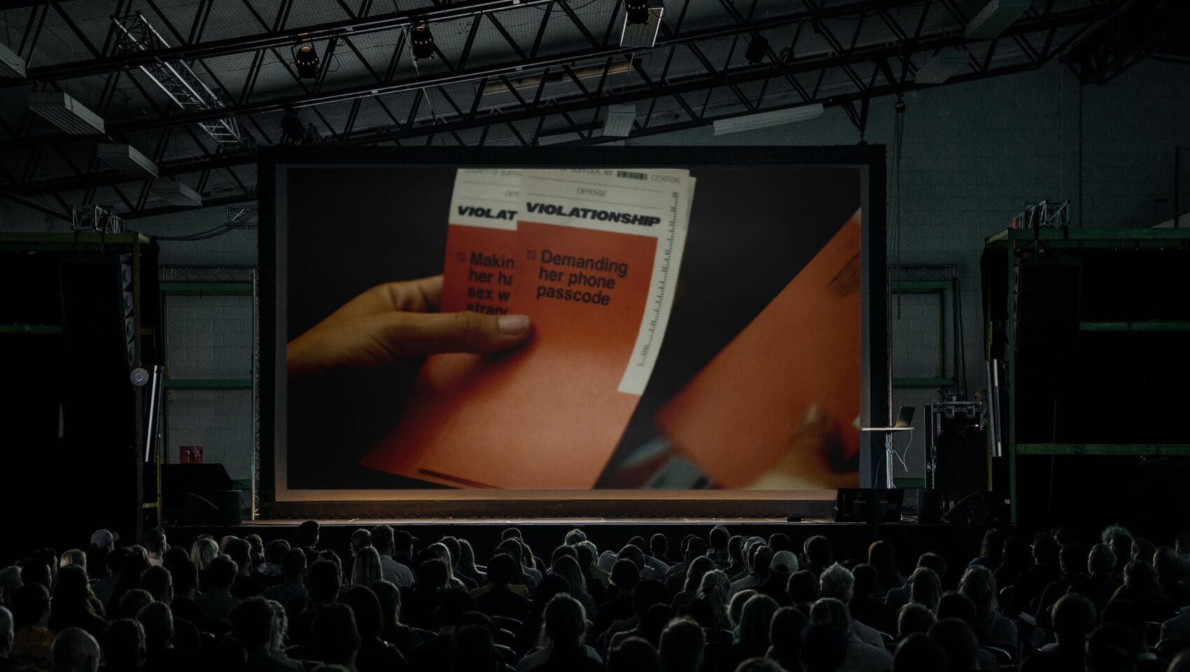 A crowded theater audience watches a large screen showing a close-up of hands holding papers labeled “VIOLATION” with listed offenses.