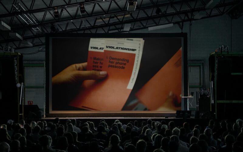 A crowded theater audience watches a large screen showing a close-up of hands holding papers labeled “VIOLATION” with listed offenses.