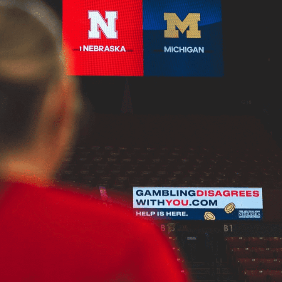 A person views a scoreboard displaying Nebraska and Michigan logos, with an anti-gambling advertisement reading "GAMBLING DISAGREES WITH YOU.COM" below.