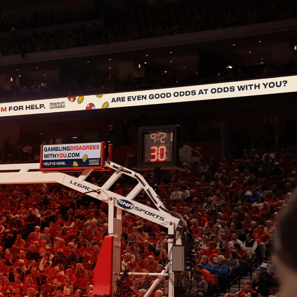A basketball court with a shot clock reading 30 and a backboard; scoreboard and gambling awareness signs are visible above a crowd of spectators.