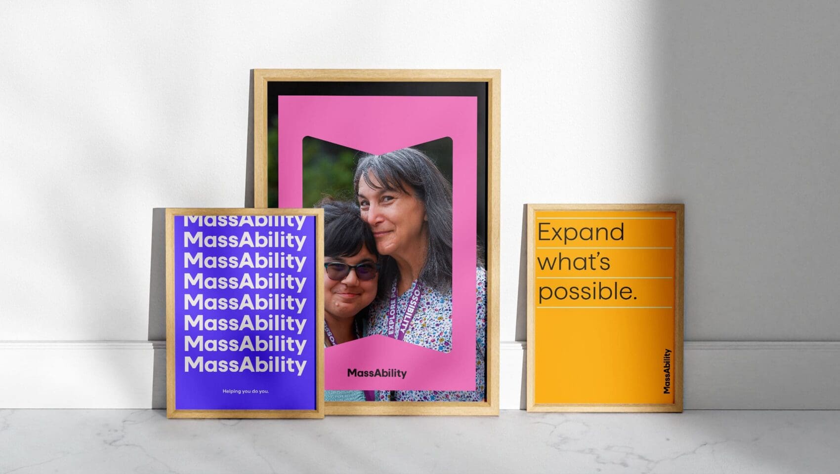 Three framed posters on a floor. Left: Repeated "MassAbility" text. Center: Two women smiling, pink background. Right: Text reads "Expand what's possible.