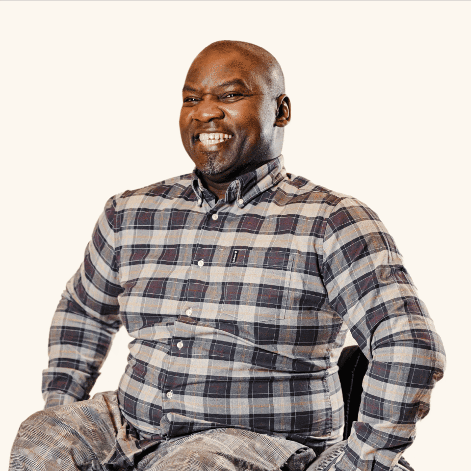 A smiling man wearing a plaid shirt sits on a chair against a plain background.