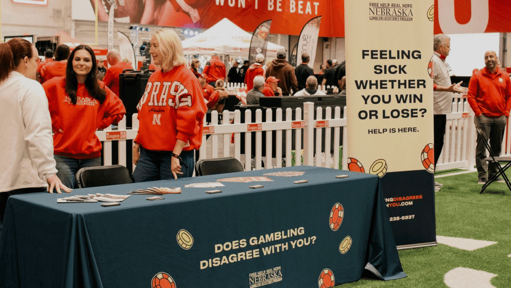 A booth with a blue tablecloth labeled "Does gambling disagree with you?" is staffed by two women at an indoor event attended by people in red clothing.