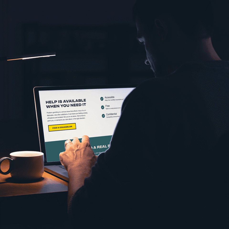 A person sits at a desk using a laptop, reading a webpage about counseling services. A cup and desk lamp are nearby, creating a dimly lit environment.