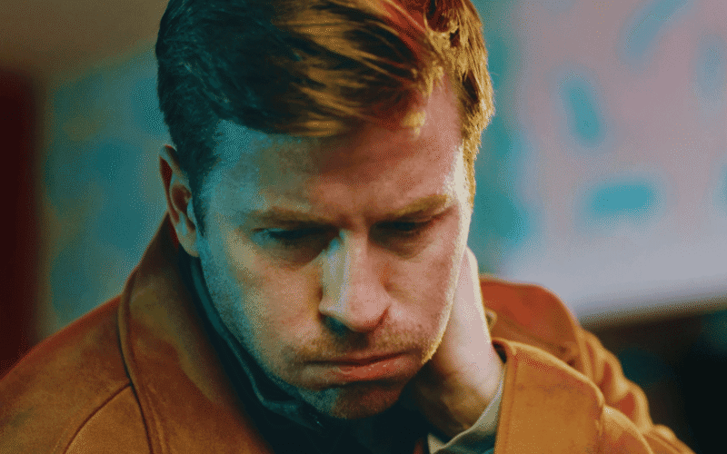 A man with light brown hair wearing a brown jacket looks down with a hand on his neck, appearing deep in thought.