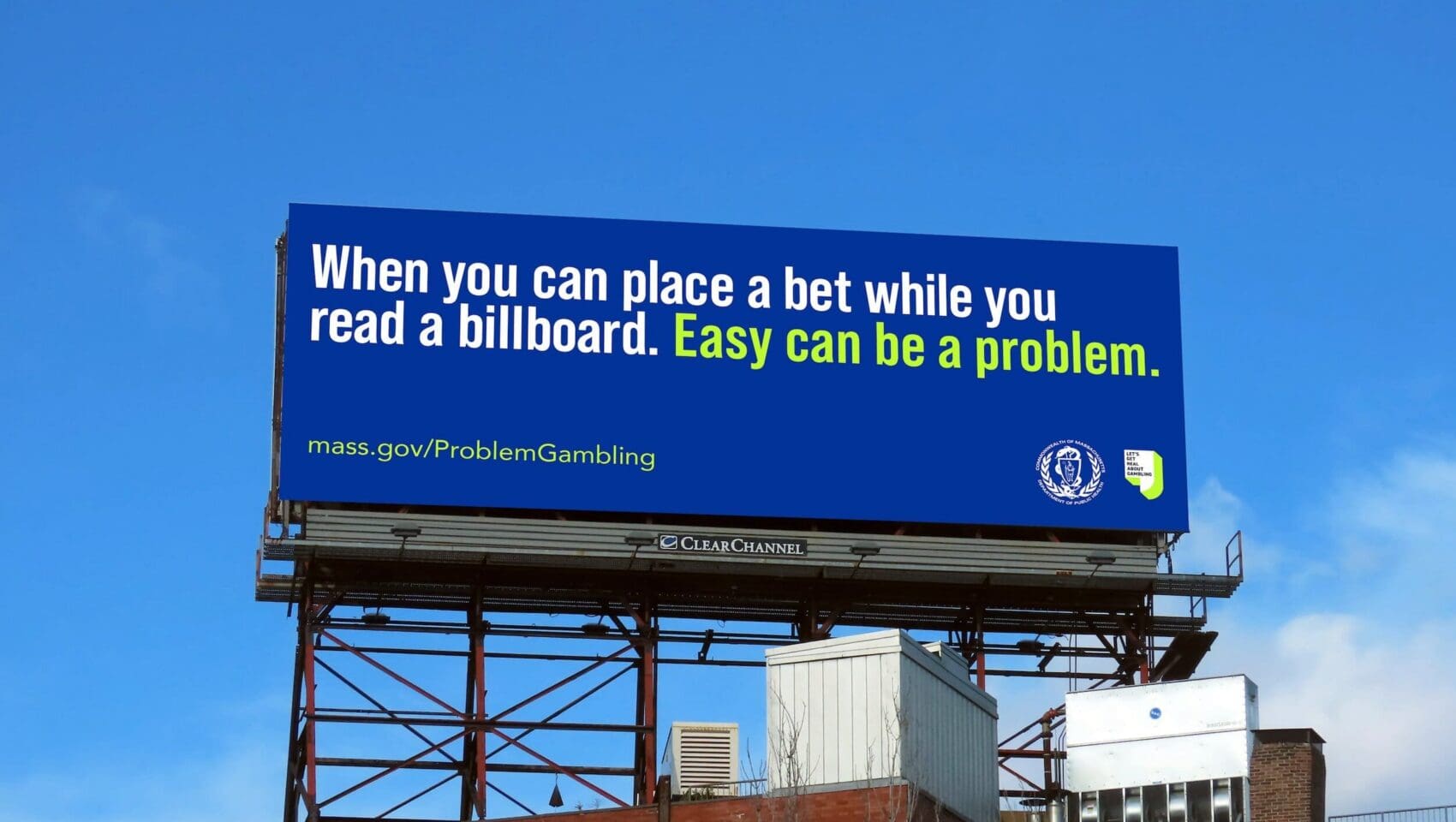 A blue billboard warns about problem gambling, stating, “Easy can be a problem,” with a website link and a state government seal, set against a clear sky above a building.