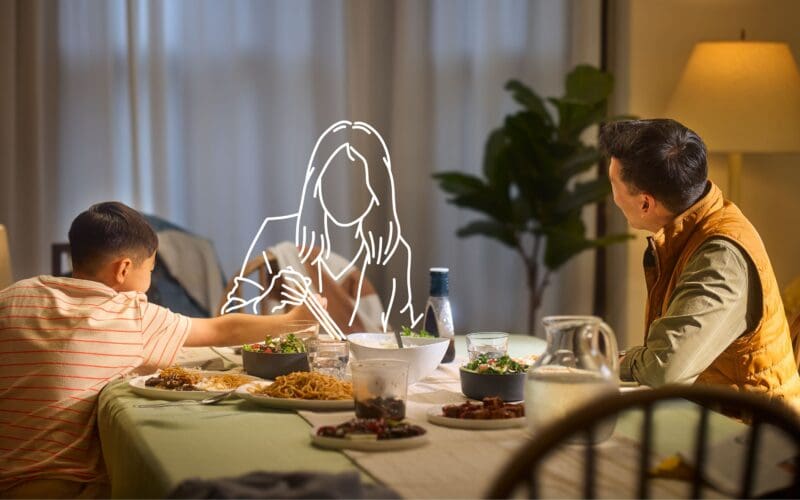 A man and a boy eat at a dinner table; an illustrated outline of a woman sits between them, suggesting her absence.