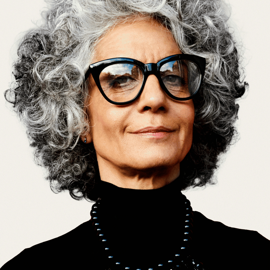 Older woman with curly gray hair, wearing large black glasses, a black turtleneck, and a beaded necklace, looks confidently at the camera against a plain background.
