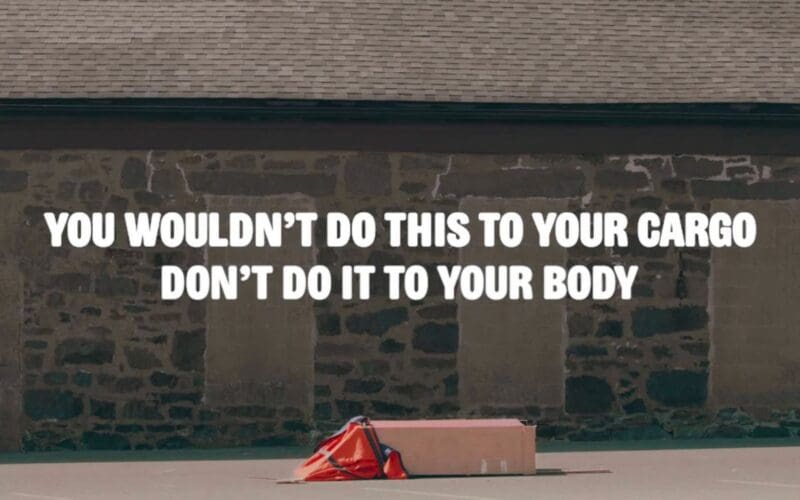 A box with an orange safety vest is dropped on pavement in front of a stone wall. Text over the image reads, “You wouldn’t do this to your cargo, don’t do it to your body.”.