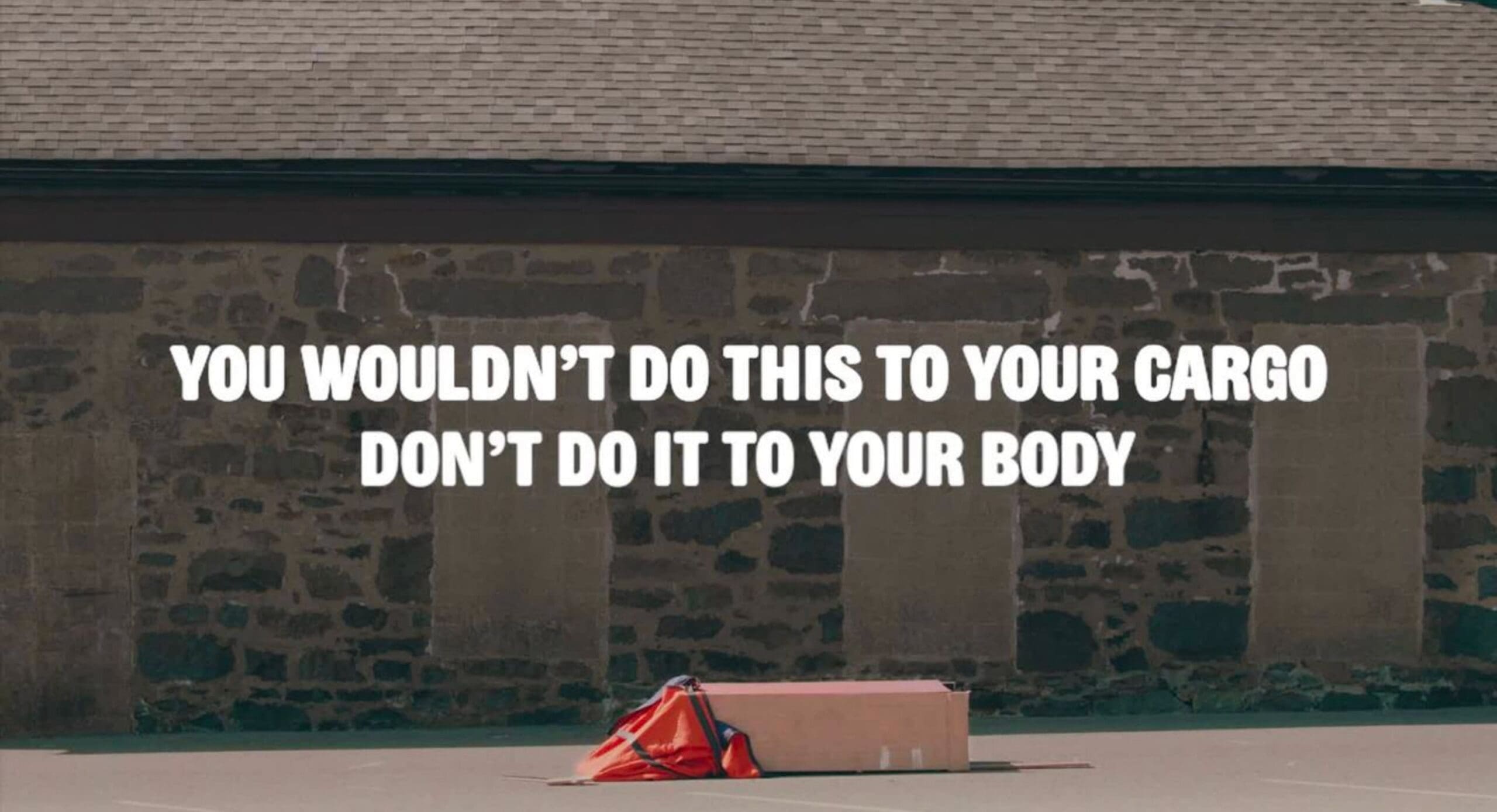 A box with an orange safety vest is dropped on pavement in front of a stone wall. Text over the image reads, “You wouldn’t do this to your cargo, don’t do it to your body.”.