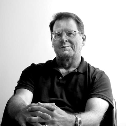 A man wearing glasses and a short-sleeved polo shirt sits with his hands clasped, looking at the camera against a plain, light background.