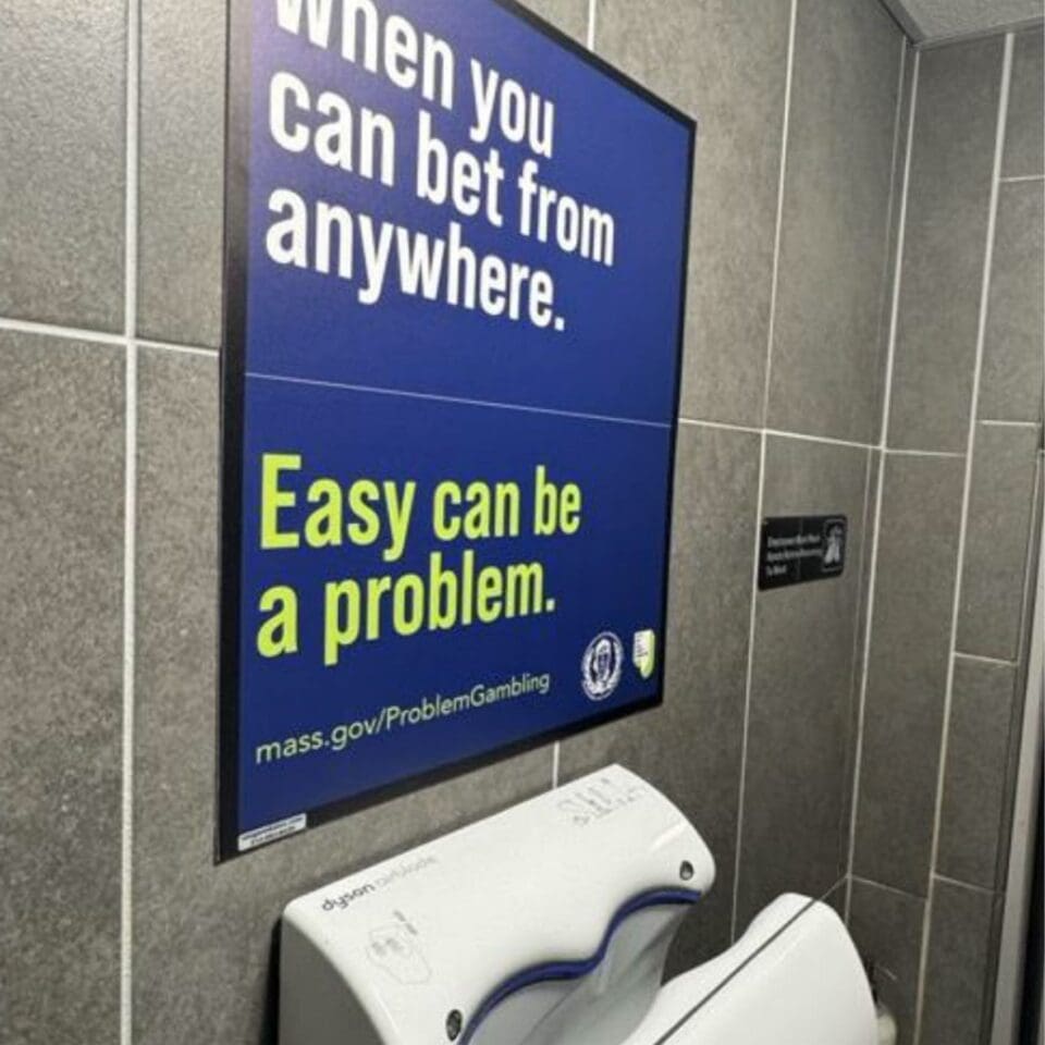 A restroom with a Dyson hand dryer and a wall sign warning about the risks of easy access to gambling, featuring a website for problem gambling help.